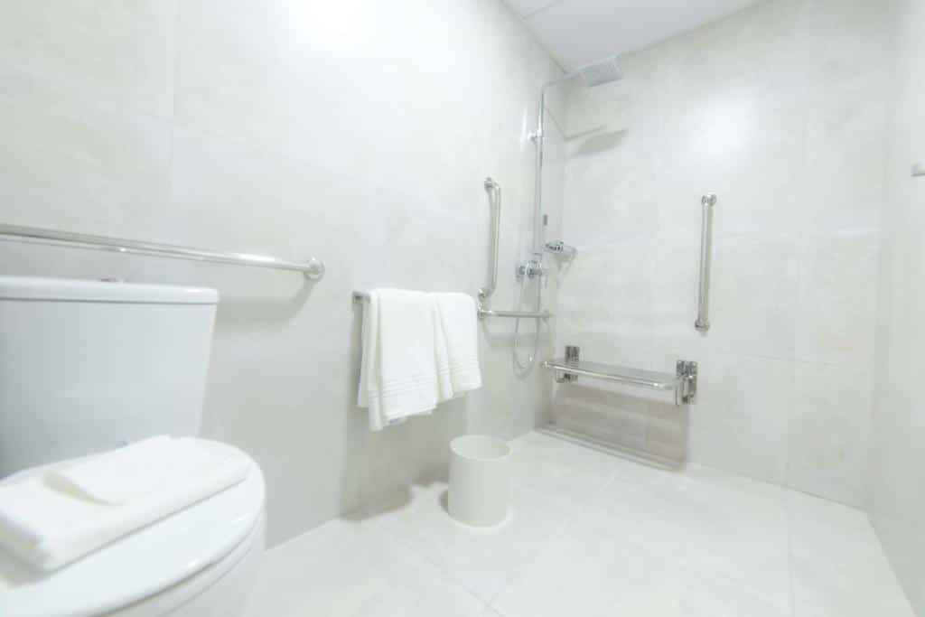 Banheiro com paredes brancas, vaso sanitário, toalhas brancas e barras de apoio para melhor acessibilidade de hóspedes com mobilidade reduzida.