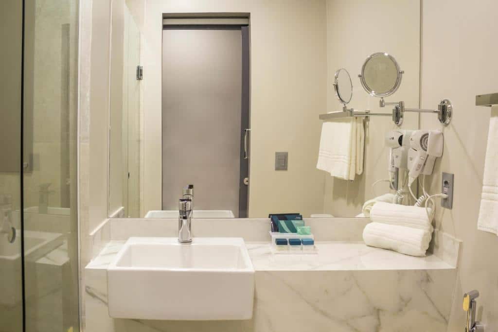 Banheiro de hotel com paredes brancas, pia em mármore branco, grande espelho e utensílios ao lado como secador de cabelo, toalha e tomada.