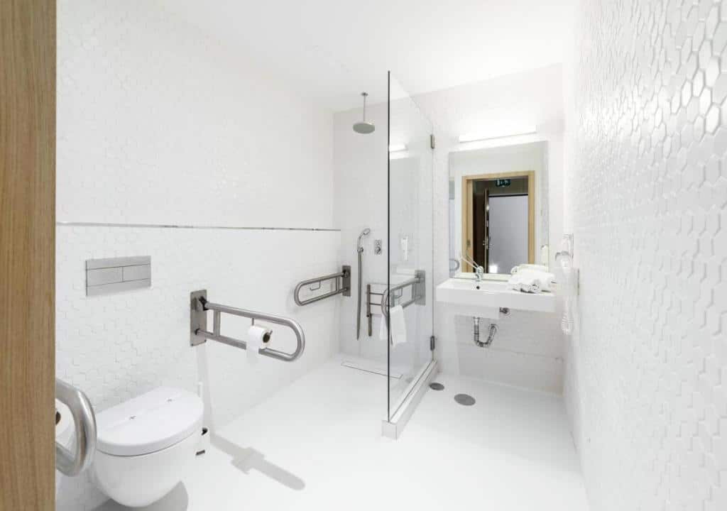 Banheiro com acessibilidade do  Moov Hotel Porto Centro com vaso sanitário do lado esquerdo com barras de segurança, logo ao fundo o chuveiro e do lado direito pia baixa.