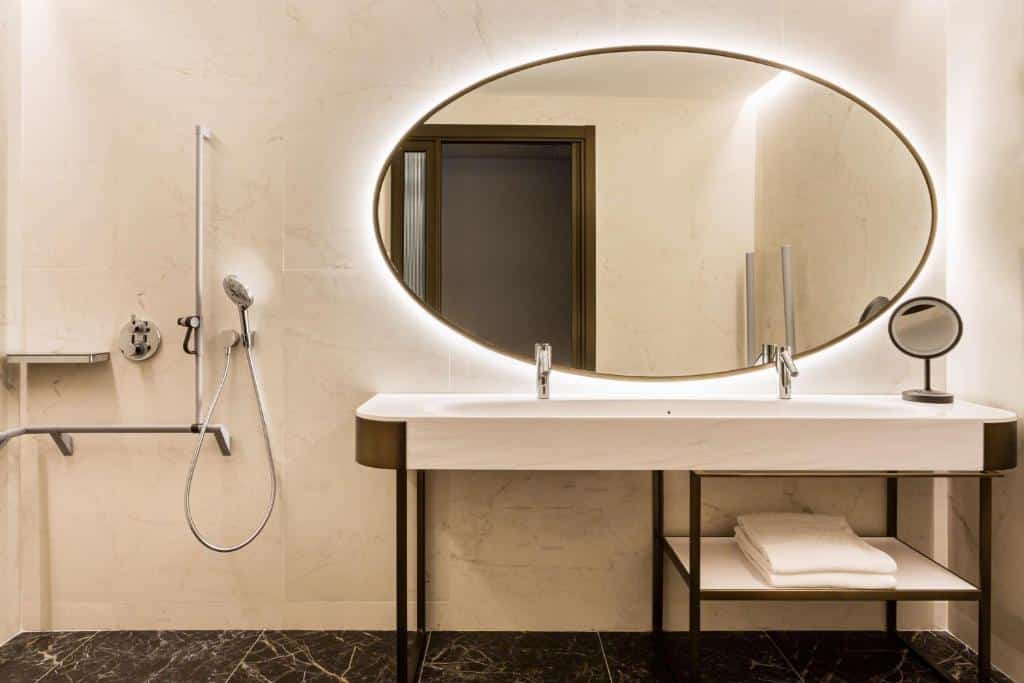 Banheiro com acessibilidade do Radisson Collection Hotel, Magdalena Plaza Sevilla. Uma barra com um chuveirinho do lado esquerdo. Do lado direito uma pia baixa e grande, com uma prateleira em baixo com toalha. Em cima da pia um espelho.