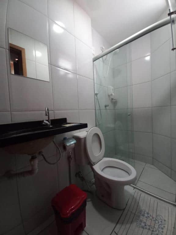 Banheiro de hotel simples com paredes brancas, pia preta, pequeno espelho, vaso sanitário e boz de vidro para o banho.