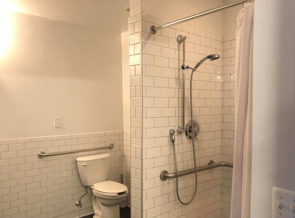 banheiro acessível do The Local NY com barras de apoio dentro do box e ao lado do vaso sanitário. O banheiro é revestido de azulejos brancos, tanto no chão quanto na parede.