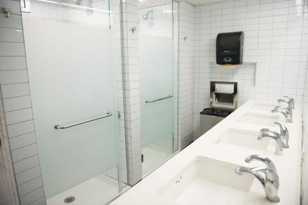 banheiro do West Side YMCA com uma bancada de pias dispostas lado a lado, em frente aos boxes de vidro dos chuveiros. O banheiro é revestido de azulejos brancos.