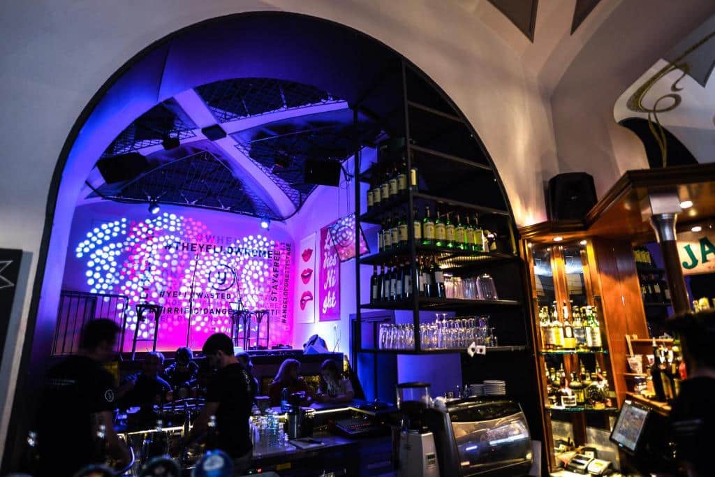 bar do YellowSquare Rome, com uma grande estante cheia de bebidas em estilo industrial, mesas com cadeiras e um grande palco com luzes roxas