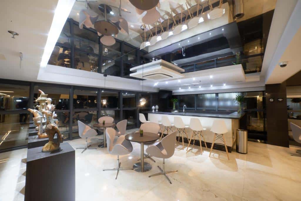 Bar de hotel com bancada preta e branca, cadeiras brancas e cinzas e teto de espelho refletindo o bar.