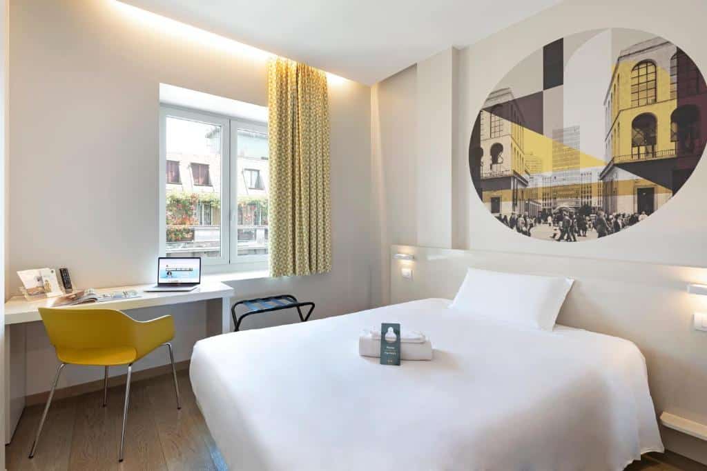 Quarto do B&B Hotel Milano Central Station, com uma janela com cortinas, uma cama de casal, uma mesa de escritório com uma cadeira, o chão imita madeira e a decoração é em preto, amarelo e branco, para representar hotéis Ibis em Milão
