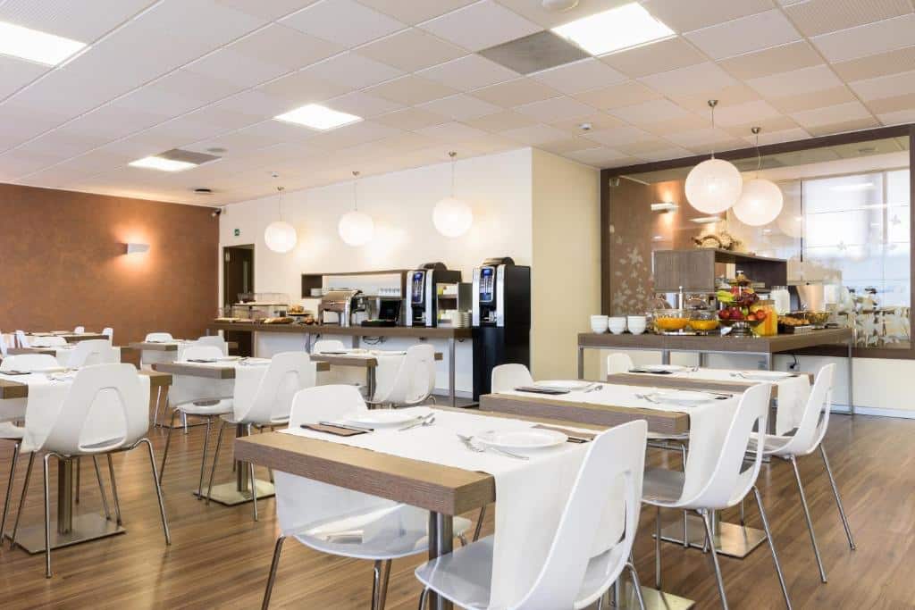 Salão de refeições do B&B Hotel Milano Portello com mesas quadradas brancas, cadeiras, e dois balcões com máquinas de café, itens de café da manhã, para representar hotéis Mercure em Milão