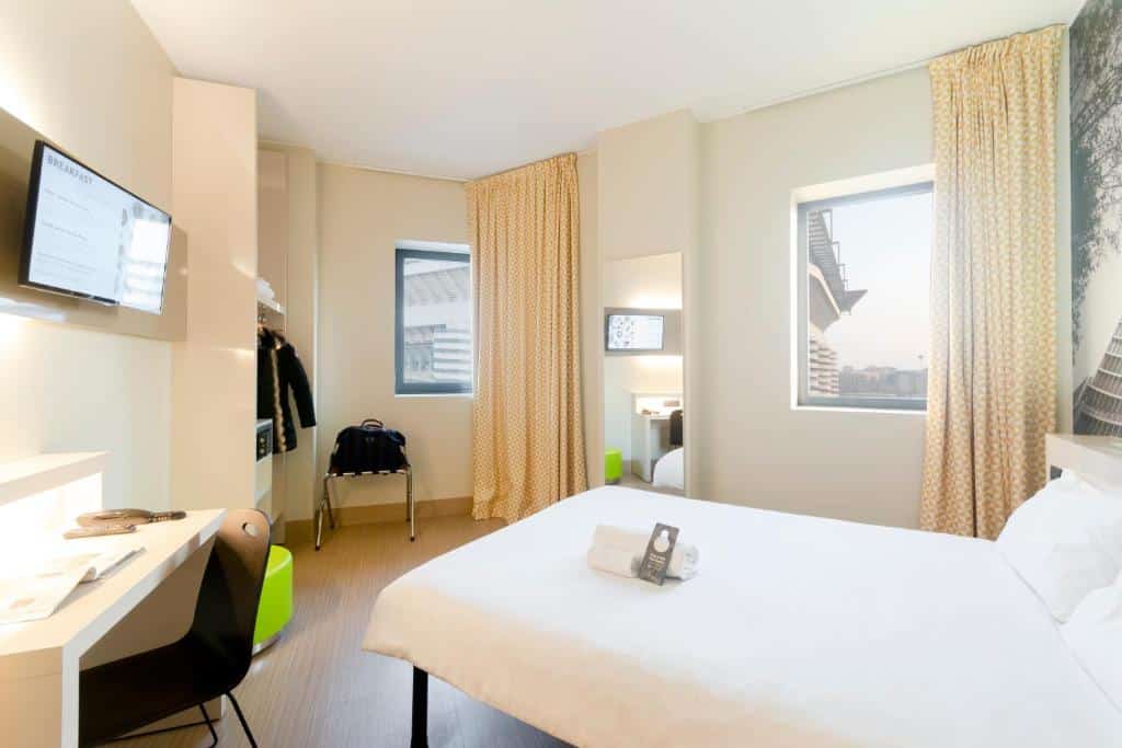 Quarto do B&B Hotel Milano San Siro com uma cama de casal, uma janela com cortinas, um espelho de corpo inteiro, um armário de conceito aberto com cabideiro, uma televisão e uma mesa de escritório com uma cadeira