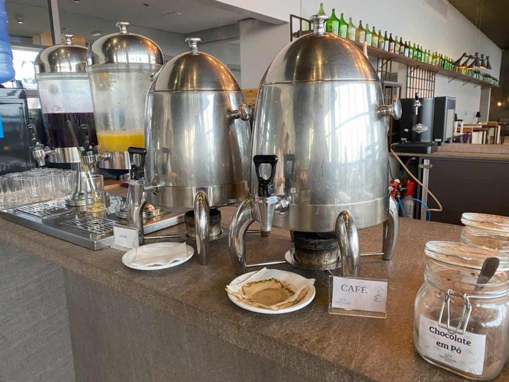 Galões com bebidas armazenadas no café da manhã do Dall'Onder Ski, sendo um com café, outro com leite, e dois com sucos, um de laranja e outro de uva, ao fundo