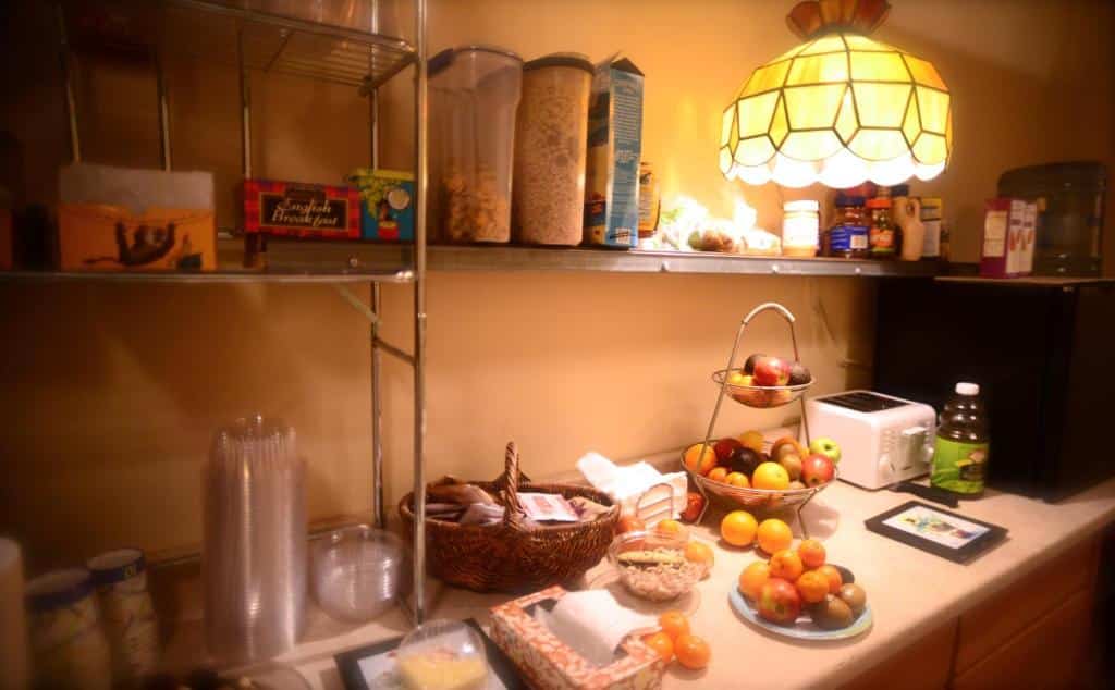 cozinha do Interfaith Retreats com um balcão repleto de alimentos, como frutas, cereais, torradeiras, utensílios de cozinha e iluminação indireta e amarelada.