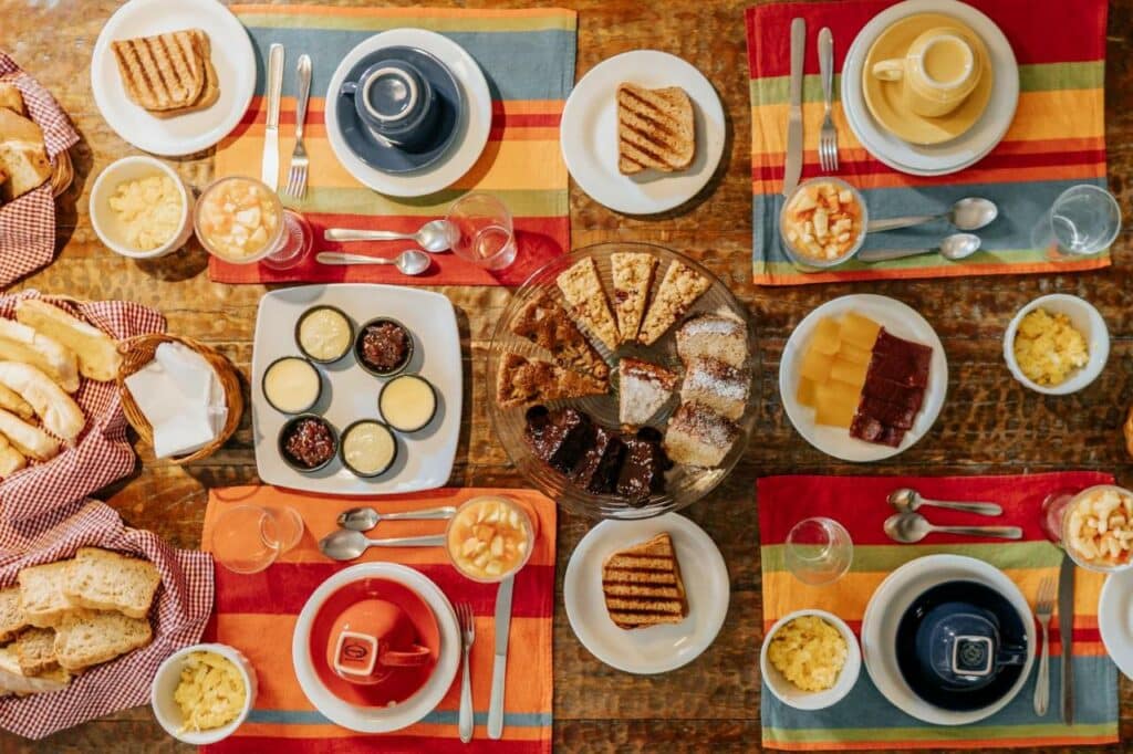 Mesa do café da manhã no Sollar dos Geranios. Uma mesa cheia de variedade e colorida, com frutas, pães, bolos, frios e xícaras.