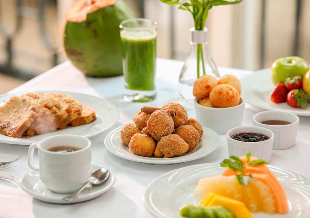 Mesa de café da manhã do Estanplaza Paulista. Na foto vemos bolinhos, pães de queijo, frutas e fatias de torradas, um café, suco verde e alguns molhos.
