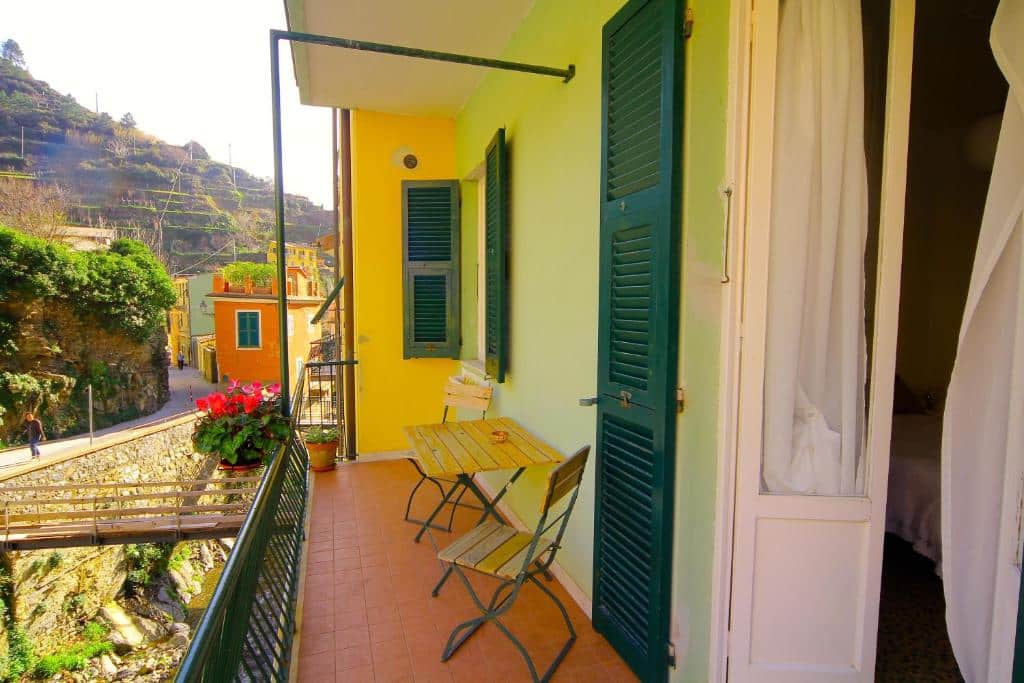 Varanda do hotel com uma porta branca que dá acesso ao quarto, janelas verdes, chão marrom, uma mesa com cadeiras de madeira e vista para a rua da cidade durante o dia, ilustrando post Hotéis em Cinque Terre.