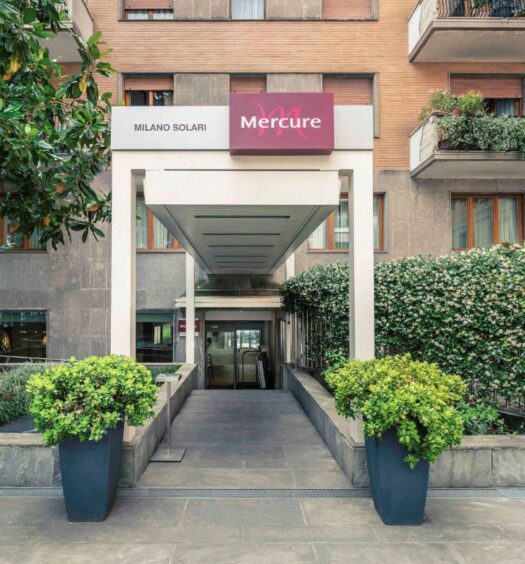 Entrada do Hotel Mercure Milano Solari com diversos vasos de plantas e arbustos criando um corredor que leva até o hall do local, para representar hotéis Mercure em Milão