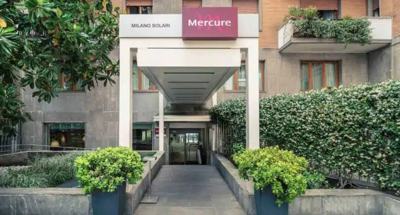 Entrada do Hotel Mercure Milano Solari com diversos vasos de plantas e arbustos criando um corredor que leva até o hall do local, para representar hotéis Mercure em Milão.