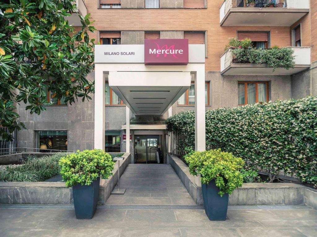 Entrada do Hotel Mercure Milano Solari com diversos vasos de plantas e arbustos criando um corredor que leva até o hall do local, para representar hotéis Mercure em Milão