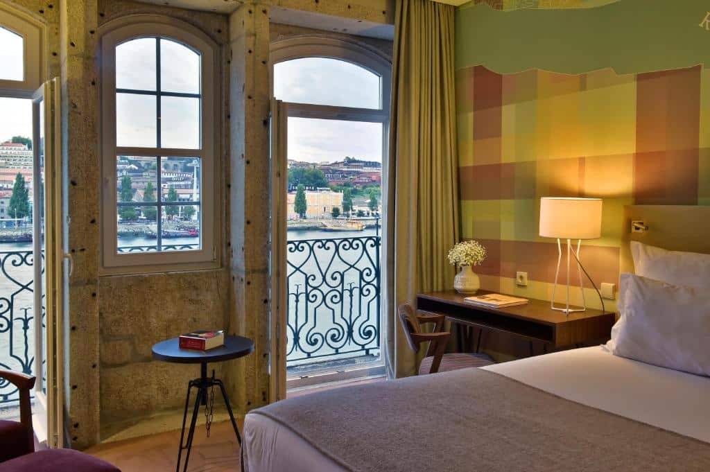 Quarto de casal do Pestana Vintage Porto Hotel & World Heritage Site com cama a frente, do lado esquerdo da cama uma mesa de trabalho e ao fundo portas amplas com vista para o Rio Douro. Representa hotéis bem localizados no Porto.