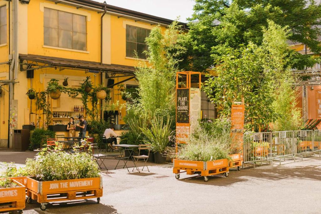 Espaço compartilhado do casaBASE com um bar, muitas árvores e plantas, além das mesas e cadeiras de madeira, para representar hostels em Milão
