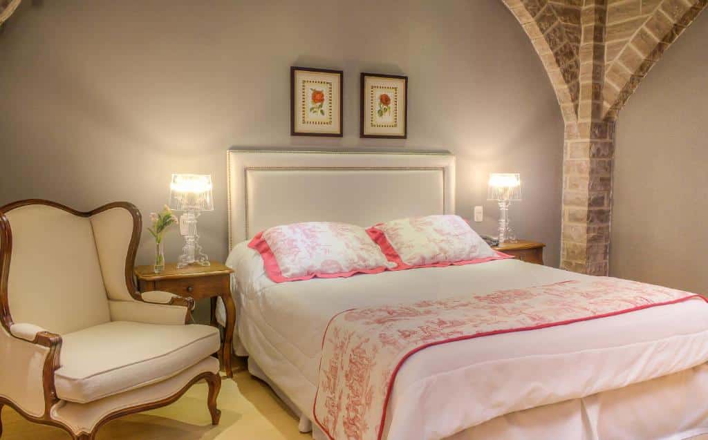 Quarto da Pousada Castelo Benvenutti, uma das opções de onde ficar no Vale dos Vinhedos, com cama de casal ampla, mesinhas de cabeceira, e uma poltrona ao lado da cama