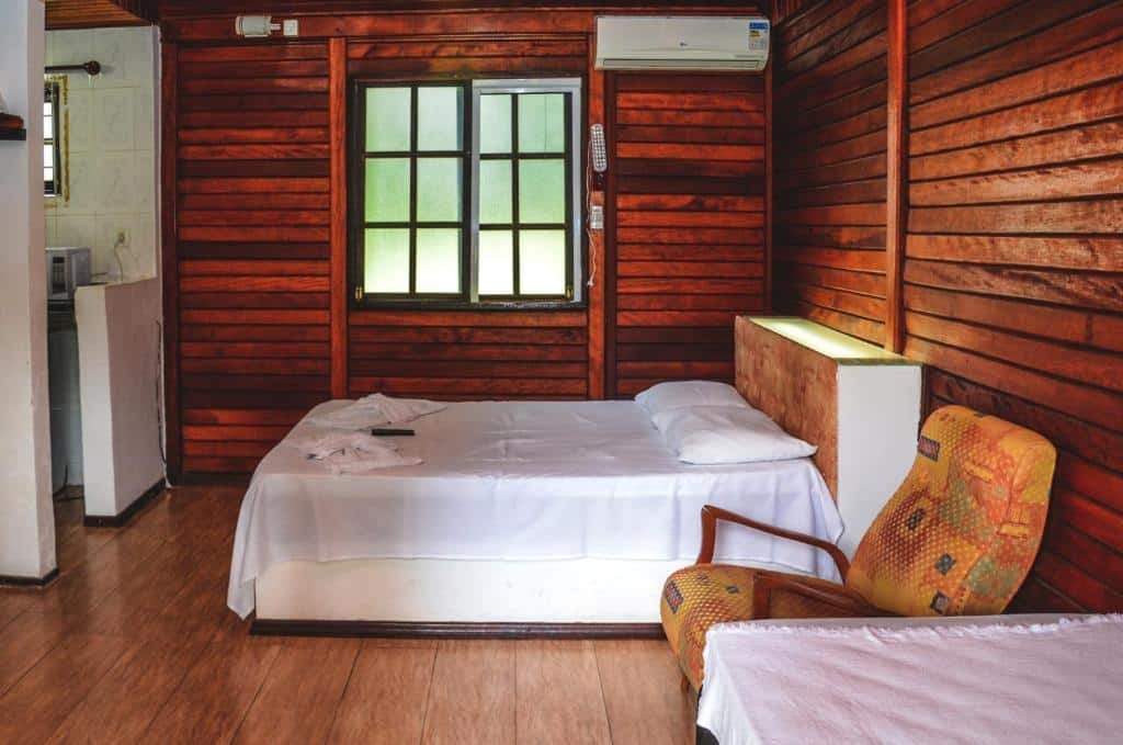 Chalé com paredes de madeira, cama de casal com lençóis brancos e poltrona estampada ao lado da cama. Imagem para ilustrar o post hotéis e pousadas em Itu.