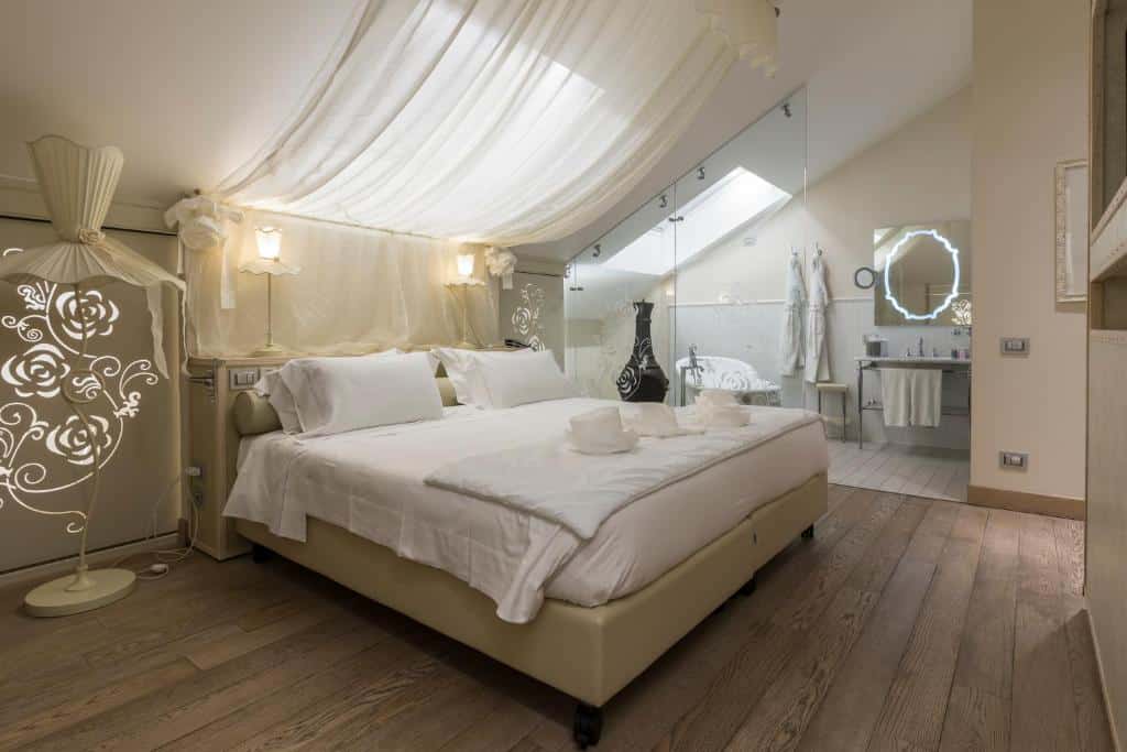 Quarto do Château Monfort - Relais & Châteaux com uma cama de casal, piso que imita madeira, uma janela em 90 graus sob a cama, há uma parede de vidro que, do outro lado, há uma banheira, uma pia ampla, um espelho e roupões