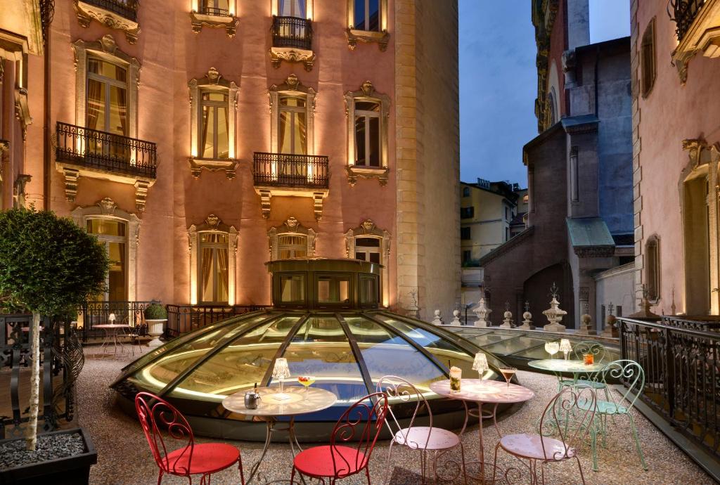Terraço do Château Monfort - Relais & Châteaux com vista para os prédios da cidade, há uma abóbada de vidro no centro do local e, nos arredores, há mesinhas e cadeiras coloridas, para representar hotéis boutique em Milão