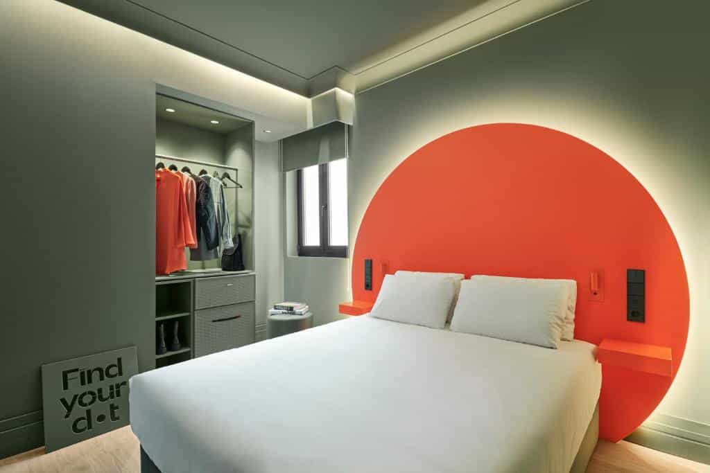 Quarto do Chic & Basic dot, uma das recomendações de hotéis baratos em Madri. Uma cama de casal tem mesinhas de cabeceira e luminárias dos dois lados. Uma ilustração de círculo coral decora a parede atrás da cama. Há um armário com roupas em cabides logo ao lado, e uma pequena janela.