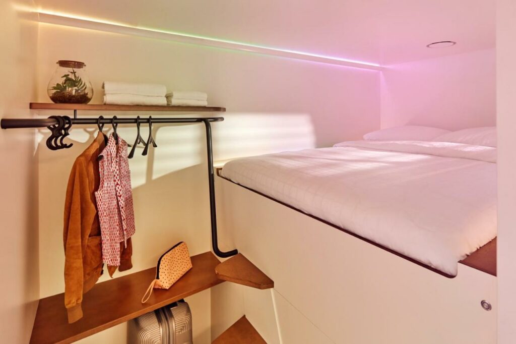 Quarto cabine do CityHub Copenhagen com uma cama de casal mais alta, duas prateleiras e uma arara com roupas. Foto para ilustrar post hotéis em Copenhague.