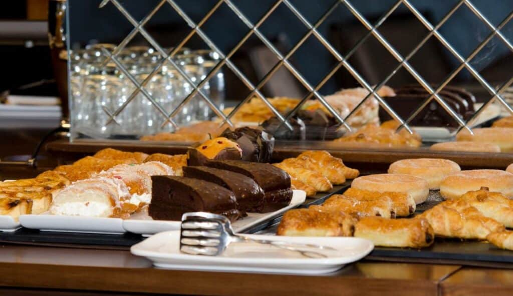 Itens na bancada de café da manhã no Claridge Madrid. Pedaços de bolo, sonhos e folhados estão dispostos com um pegador num prato bem em frente.