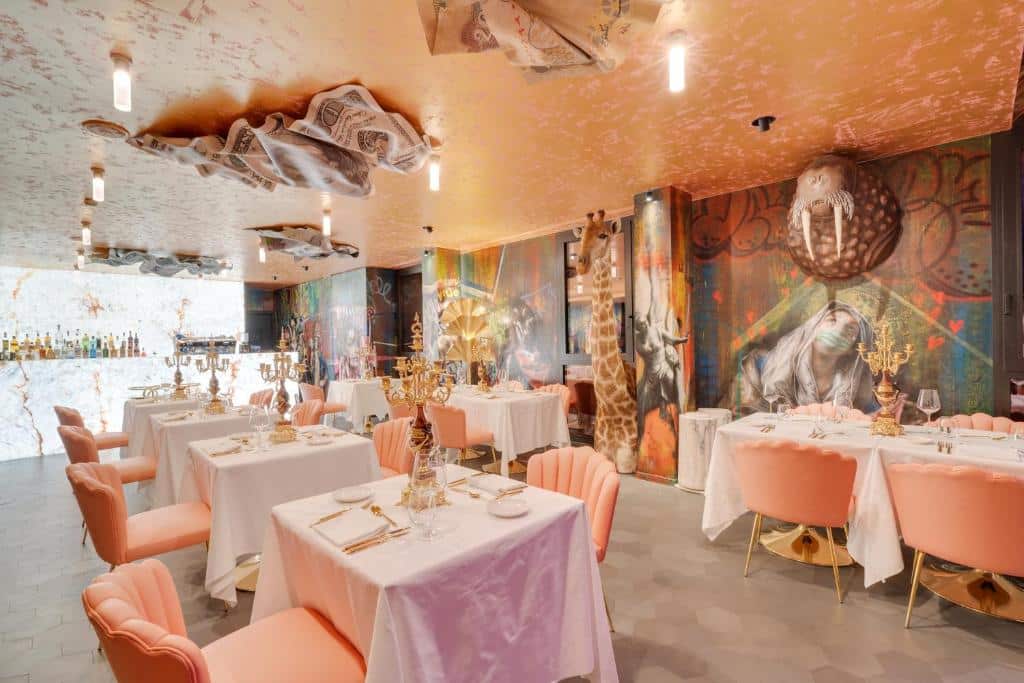 Salão de refeições do Collini Rooms, tudo decorado em um estilo de fantasia, no teto há falsas folhas de jornal presas, as mesas são quadradas e as cadeiras estofadas em tom de salmão, as paredes são pintadas e tem esculturas de animais presos