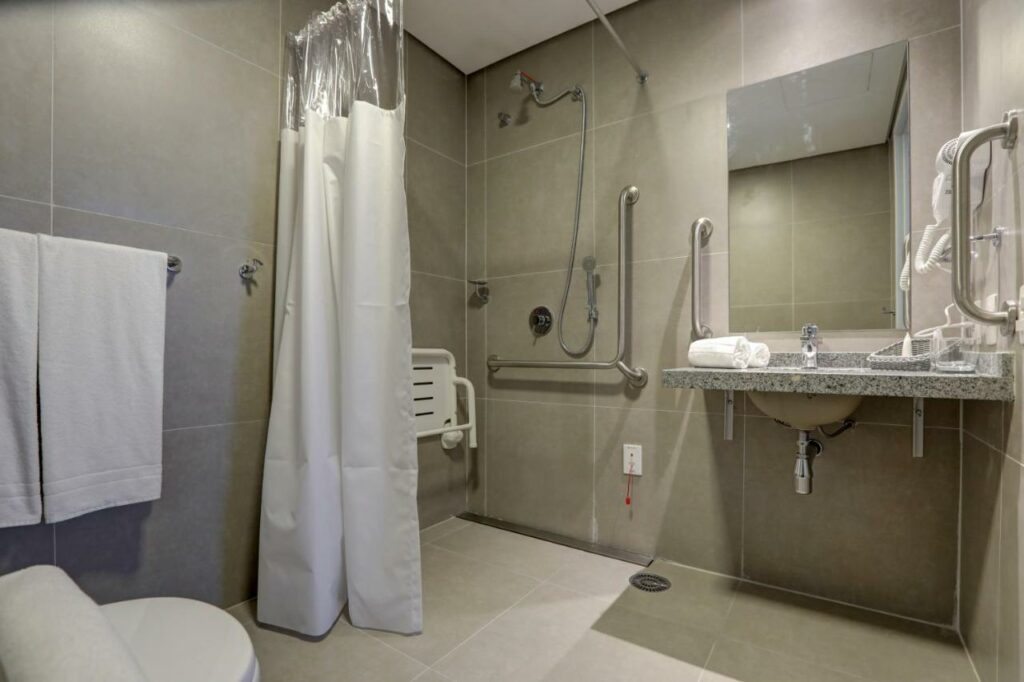 Banheiro acessível para PcDs no Comfort Hotel Guarulhos Aeroporto, com assento no chuveiro, cordão de emergência, pia rebaixada e barras de apoio