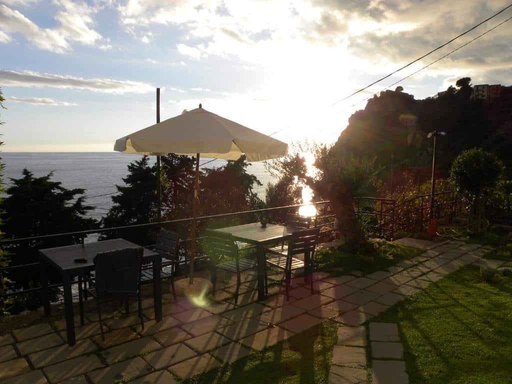 Duas mesas com cadeiras, um guarda-sol, várias árvores em volta, gramado verde e uma vista com sol para o mar da cidade, ilustrando post Hotéis em Cinque Terre.