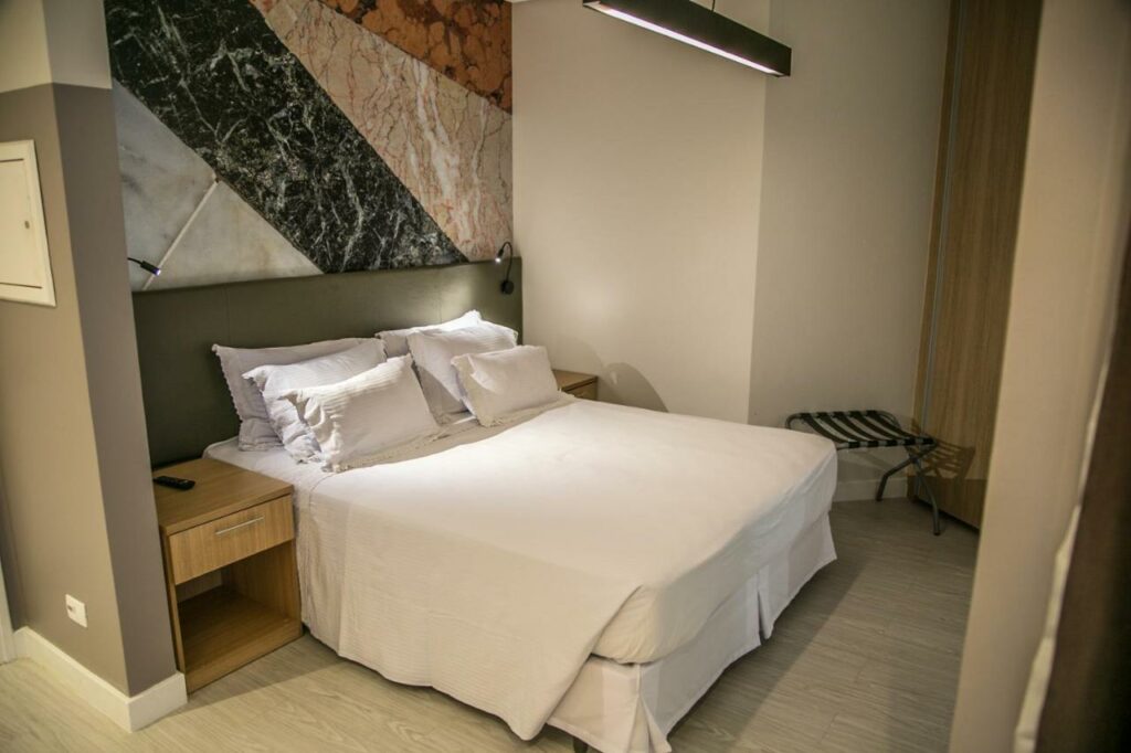 Quarto do Cosmopolitan Praia Flat com uma cama de casal e uma mesinha. A parede atrás da cama é bem decorada com uma pintura. Foto para ilustrar post sobre pousadas em Santos.