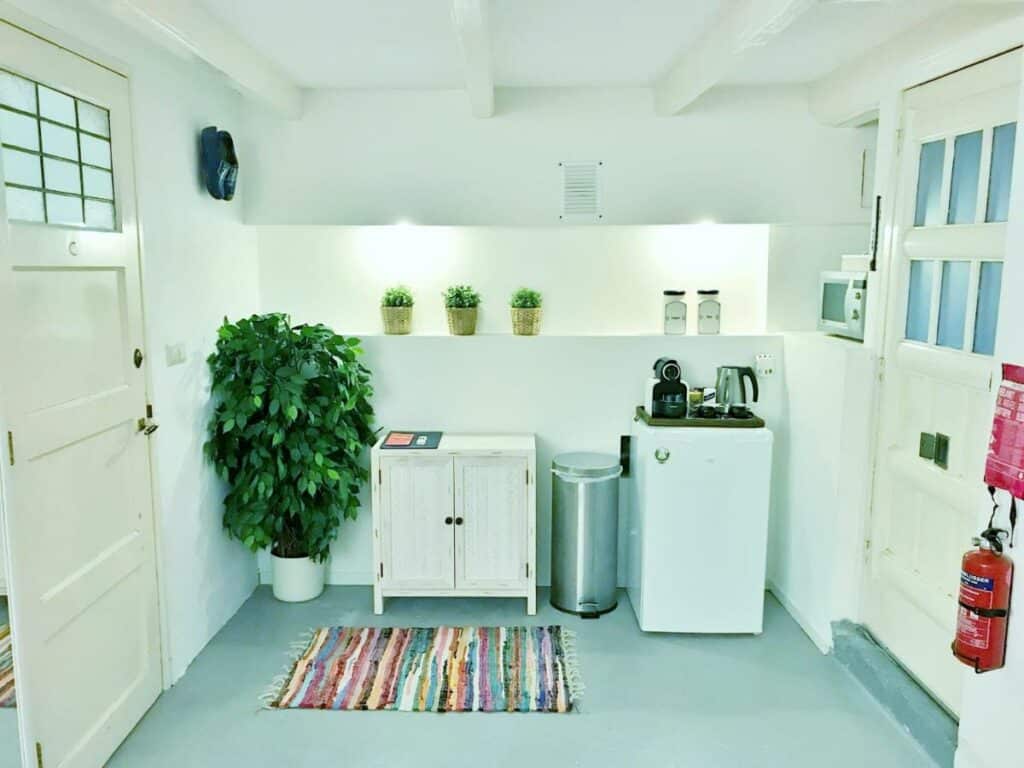 Cozinha compacta do Amsterdam Home. com um móvel com duas portas branco, um lixo de alumínio e, ao lado, um frigbar com utensílios em cima. Há vasos de flores e um tapete pequeno colorido enfeitando o ambiente