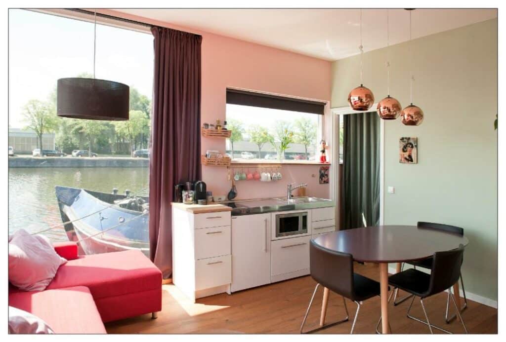 Interior do Private guesthouse BnB The Waterhouse houseboat, de 35 m², com uma mesa redonda preta com duas cadeiras, uma pia pequena ao fundo com ármário em baixo, no lado esquerdo tem um sofá vermelho e ao lado uma porta de vidro mostrando um rio. Representa airbnb em Amsterdam