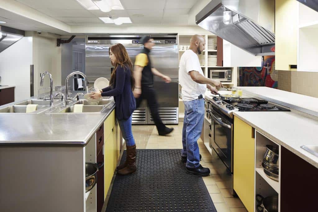 Uma imagem de uma cozinha no HI New York City Hostel em Nova York. A cozinha é espaçosa e bem iluminada, com uma variedade de equipamentos, incluindo uma geladeira, um fogão, um forno, um micro-ondas, duas pias para lavar louça diversas panelas.