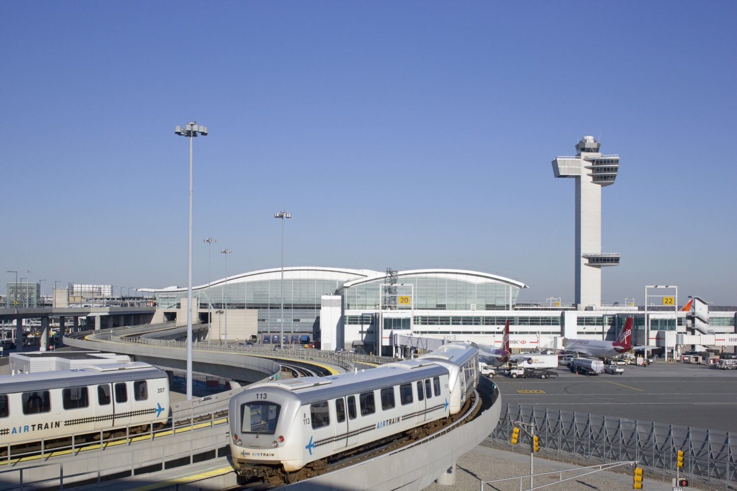 vista da área externa do aeroporto internacional JFK de Nova York mostrando duas trilhas com carros de metrô à esquerda da imagem e à direita, alguns aviões taxiando na pista de pouso. Ao fundo é possível ver uma torre de controle sobre um céu azul e sem nuvens