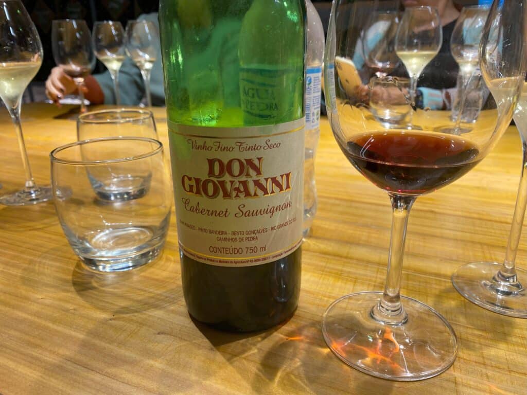Close em garrafa de vinho fino tinto seco, de uma das opções de vinícolas no Vale dos Vinhedos, a Don Giovanni, da safra de 1996, variedade Cabernet Sauvignon, com taça ao lado, sobre mesa de madeira
