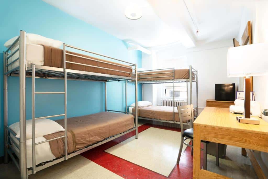 dormitório compartilhado do West Side YMCA, um dos hostels em Nova York, com paredes azuis e brancas, duas beliches de metal com lençóis brancos e marrons, e uma mesa de madeira no canto direito da imagem para quem precisa trabalhar.