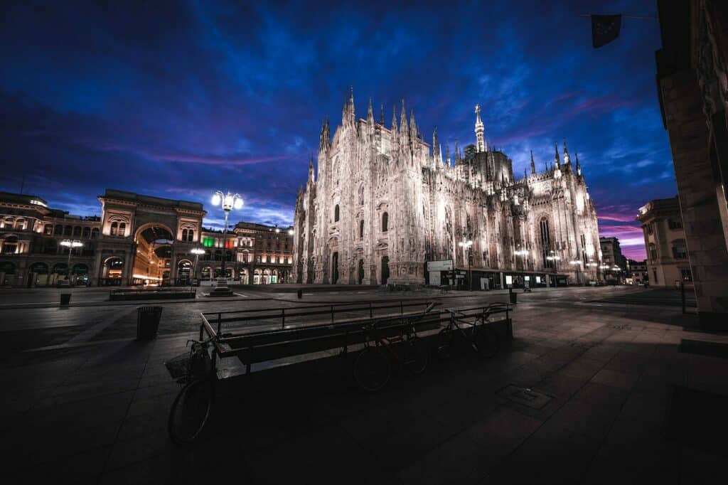Catedral de Milão em estilo gótico iluminada de noite, o pátio que forma a Praça Duomo está vazio e é possível ver outros prédios históricos no local