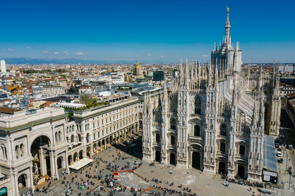 Vista aérea da Praça Duomo com foco na Catedral de Milão em estilo gótico, há um pátio na frente da igreja com muitas pessoas andando, para representar contratar seguro viagem é obrigatório