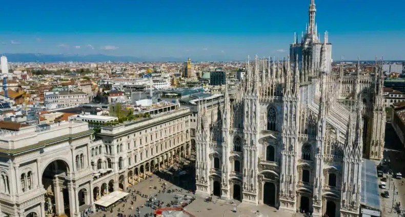 Vista aérea da Praça Duomo com foco na Catedral de Milão em estilo gótico, há um pátio na frente da igreja com muitas pessoas andando, para representar onde ficar em Milão