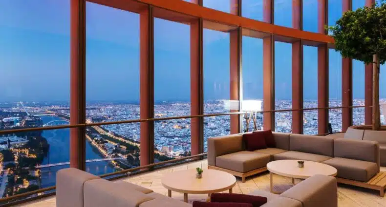 Dois sofás em formato grandes e cinzas com duas mesas de centro, atrás o hotel de vidro com vista de 360° da cidade, no Hotel Eurostars Torre Sevilla, para representar hotéis em Sevilha.
