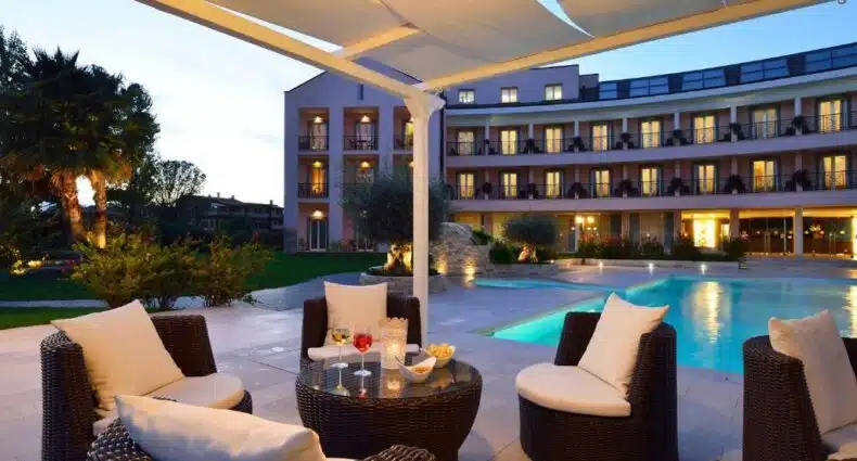 área externa do Hotel Isola Sacra Rome Airport, um dos hotéis perto do aeroporto de Roma, com piscina, fachada atrás ao entardecer com diversas luzinhas e, à frente, um gazebo com poltronas e mesinhas com bebidas postas