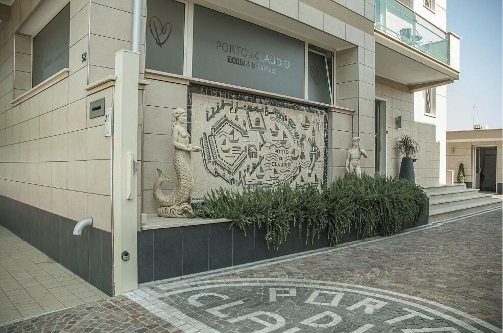 fachada do Porto Di Claudio, com mosaico escrito o nome do hotel no chão e também na parede, com esculturas na frente, sendo uma sereia e um herói clássico romano e vidros