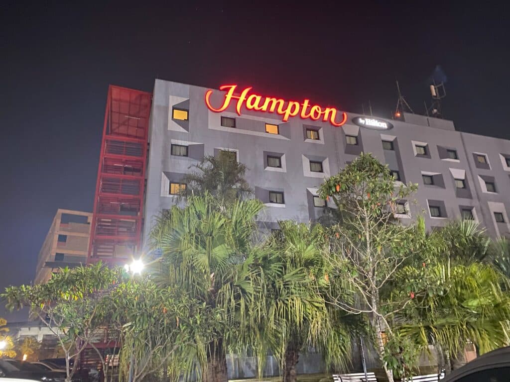 Fachada do Hampton inn by Hilton, um dos hotéis perto do aeroporto de Guarulhos, com letreiro do nome do hotel iluminado à noite, e árvores tampando parte da fachada, que é vista de baixo para cima