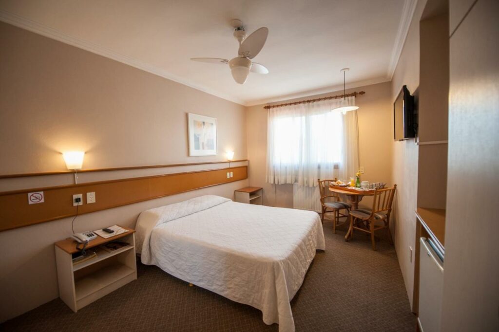 Quarto do Farina Park Hotel, com cama de casal, mesa de cabeceira, mesa redonda com duas cadeiras no canto, além de ventilador de teto, TV na parede e frigobar