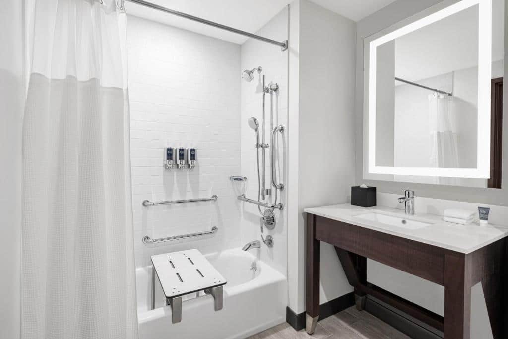 banheiro do Four Points by Sheraton Flushing mostrando adaptações para pessoas com mobilidade reduzida, tais como barras dentro do box, com uma banheira de chão, um banco de banho e uma pia vazada com um espelho amplo e quadrado.