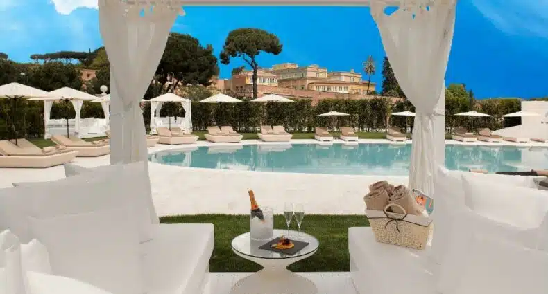 gazebo na beira da piscina com sofás e mesinha brancos com espumante servido em frente à piscina com mais mesinhas ao fundo e vista de monumentos da cidade no Villa Agrippina Gran Meliá, um dos hotéis românticos em Roma