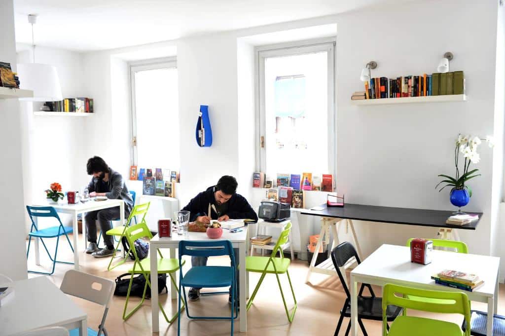 Espaço compartilhado do Gogol'Ostello & Caffè Letterario com janelas, livros, mesas e cadeiras, há dois rapazes em mesas diferentes fazendo uso do local